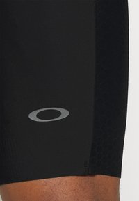 Shorts de sport noirs en tissu lisse avec un panneau latéral texturé présentant un motif en nid d'abeille. Un logo circulaire argenté est affiché.