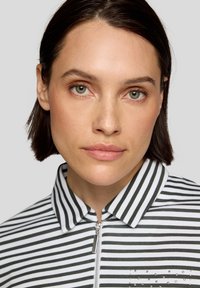 Reißverschluss-Oberteil mit Kragen, das schwarze und weiße vertikale Streifen aufweist, mit einer strukturierten Fronttasche, die mit kleinen Nieten verziert ist. Glatter Stoffabschluss.