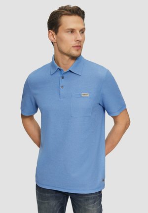 Hellblaues Poloshirt aus strukturiertem Stoff, mit Kragen, drei Knöpfen und einer Brusttasche mit Label. Kombiniert mit dunkler Jeans.