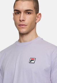 Camiseta de color púrpura claro hecha de algodón, con un logotipo bordado rectangular en rojo, negro y blanco en el lado izquierdo del pecho.