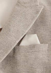 Béžový texturovaný klopový golier saka s krémovo bielym vreckovým obrúskom starostlivo zloženým vo vrecku na hrudi.