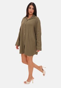 Olijfgroene oversized jurk met een textuur van gekreukeld stof. Voorzien van een knoopsluiting, lange mouwen en zijsplitten. gedragen met beige hakken.