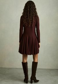Bordo spalvos ilgos rankovės suknelė su platėjančia sijonu, tekstūruota medžiaga ir raukšlėmis, derinama su ruda zomšine, iki kelių siekiančia avalyne.