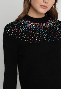 Schwarzer Pullover mit tailliertem Design, ausgestattet mit bunten Paillettenverzierungen an den Schultern, die Textur und Kontrast hinzufügen.