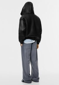 Svart huva jacka med plyschfoder, kombinerad med lösa grå byxor med subtila mönster och sidofickor.