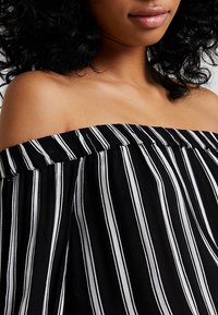 Blouse décolletée mettant en avant un tissu noir avec des rayures verticales blanches, un col froncé et un matériau léger et texturé.