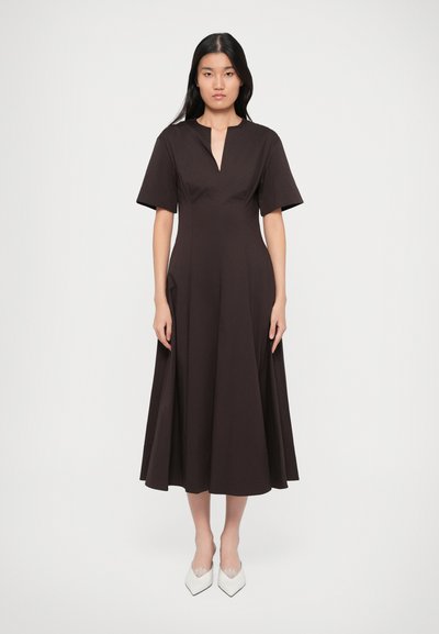 Braunes knielanges Kleid mit kurzen Ärmeln, tiefem V-Ausschnitt und figurbetonter Oberteil, das in einen voluminösen Rock übergeht. Tragen dazu weiße spitze Schuhe.