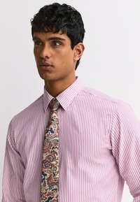 Homme aux cheveux noirs, courts et bouclés, portant une chemise à rayures roses et blanches et une cravate multicolore à motif cachemire, regardant sur le côté.