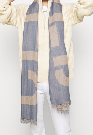 Personne portant un pull en laine crème et un pantalon blanc avec une longue écharpe bleue et beige à franges drapée autour du cou.