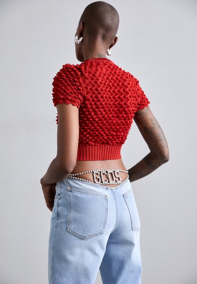 Top corto de punto texturizado rojo con mangas abullonadas, combinado con unos vaqueros de tiro alto en azul claro. Los vaqueros tienen un cinturón decorativo de perlas con letras.