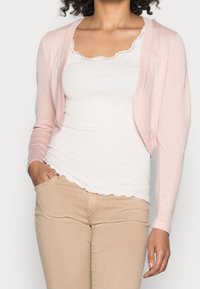 Ljusrosa cardigan med långa, åtsittande ärmar och öppen front, bärs över en vit ribbad linne och bruna byxor, med en mjuk textur.