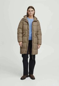 Manteau duvet marron, avec une fermeture éclair et des boutons-pression, des poches latérales et une capuche. Porté sur un haut en tricot bleu clair et un pantalon noir.