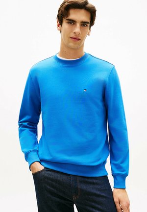 Giovane uomo che indossa una felpa girocollo azzurra con un piccolo logo sul petto e jeans in denim scuro, in piedi con una mano in tasca.