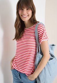 Donna sorridente con indosso una maglietta a righe rosse e bianche e jeans blu, con le mani nelle tasche, che porta una borsa tote di denim azzurro sulla spalla.