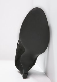 Černá semišová kotníková bota s špičatou špičkou, štíhlým podpatkem a hladkou podrážkou, umístěná podrážkou nahoru proti bílým rohovým zdím.