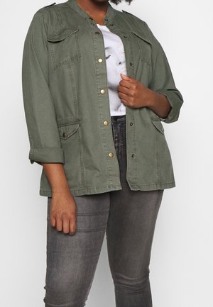 Person iført en åben olivengrøn jakke med knapper og klaplommer, en hvid t-shirt med grafik og grå tætsiddende jeans.