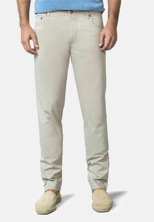 Man die lichtbeige casual broek, blauw overhemd en beige instapschoenen draagt, staand tegen een effen achtergrond.