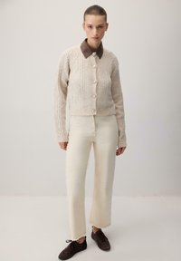 Cardigan crem cu model în cablu, cu nasturi mari și guler maro, asortat cu pantaloni crem și adidași maro închis.