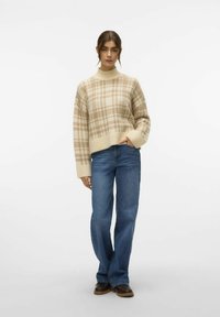 Sweter w kratę w kolorze kremowym i beżowym, z wysoką szyją i ściągaczem, zestawiony z niebieskimi spodniami o wysokim stanie i ciemnobrązowymi butami.