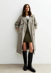 Abrigo trench de cuadros en beige y marrón, llevado sobre un vestido verde con un cuello blanco, combinado con botas altas negras. Textura suave, corte ajustado.