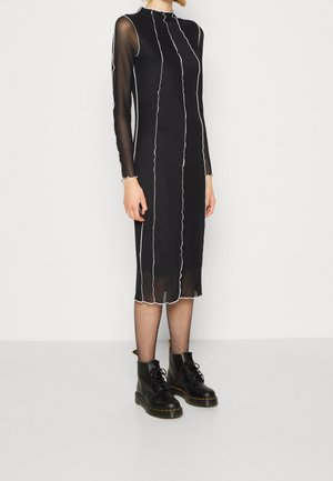 Persona que lleva un vestido midi negro con mangas largas translúcidas y costuras blancas verticales, combinado con medias de red y botas negras con cordones.