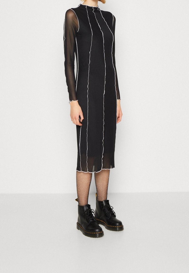 Personne portant une robe midi noire avec des manches longues transparentes et des coutures blanches verticales, associée à des collants en résille et des bottes noires à lacets.