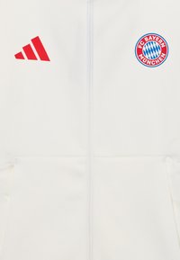 Hvit zip-up jakke med rødt Adidas-logo og FC Bayern München sirkulært merke. Glatt stoff med strukturert design og kengurulommer.