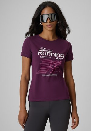 Kobieta o długich czarnych włosach, nosząca za dużą, lustrzaną sportową parę okularów przeciwsłonecznych, fioletową koszulkę "Afterlight Running Tour" i ciemne legginsy.