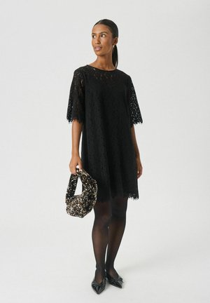 Schwarzes Spitzenkleid mit kurzen Ärmeln, locker sitzend und mit gewelltem Saum. Die Modells hält eine schwarz-goldene verzierten Tasche. Schwarze Strumpfhosen und Schuhe.