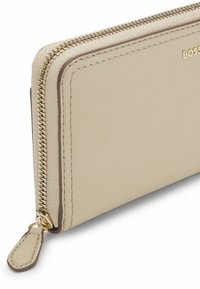 Beżowy skórzany portfel z złotym zamkiem błyskawicznym i okuciami. Charakteryzuje się eleganckim designem, gładką fakturą oraz ozdobnym przeszyciem na brzegach.