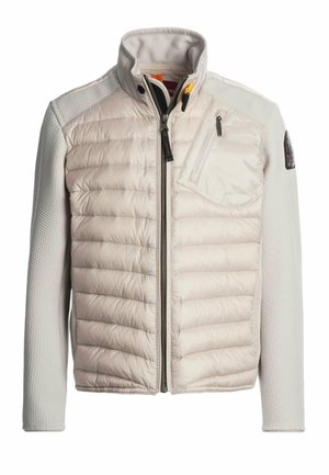 Veste matelassée beige avec manches longues texturées, col haut, fermeture éclair à l'avant et poche poitrine zippée sur le côté gauche.