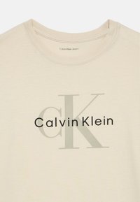 Beige pamut póló kerek nyakkivágással, nagy "Calvin Klein" logóval sötét- és világosszürkében. Puha tapintás, minimalista design.