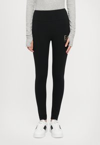 Zwarte leggings met een hoge tailleband, gemaakt van rekbaar materiaal. Heeft een klein gouden logo aan de zijkant en een gladde textuur. Gepaarde met witte sneakers.