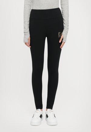 Svart leggings med høy midje, laget av elastisk materiale. Har en liten gull-logo på siden og en glatt tekstur. Kombinert med hvite sneakers.