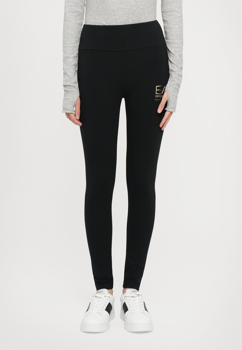 Svart leggings med høy midje, laget av elastisk materiale. Har en liten gull-logo på siden og en glatt tekstur. Kombinert med hvite sneakers.