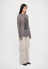 Femeie cu păr lung și închis la culoare, purtând un pulover gri transparent cu un design floral și pantaloni cargo bej cu croi larg, stând pe un fundal alb.