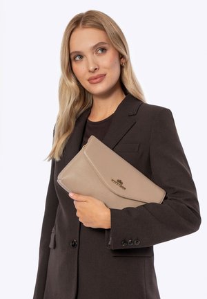 Blonde vrouw met een donker colbert houdt een beige leren clutch tas vast met een gouden "Wittchen" logo tegen een witte achtergrond.
