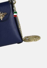 Sac en cuir bleu marine avec un emblème en forme d'abeille dorée, une bandoulière en chaîne dorée et un petit charme en forme de drapeau italien accroché. Design structuré, surface texturée.