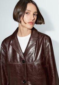 Giubbotto di pelle marrone con una texture liscia, colletto a punta, bottoni frontali e dettagli di cucitura in vista, abbinato a una camicia bianca.