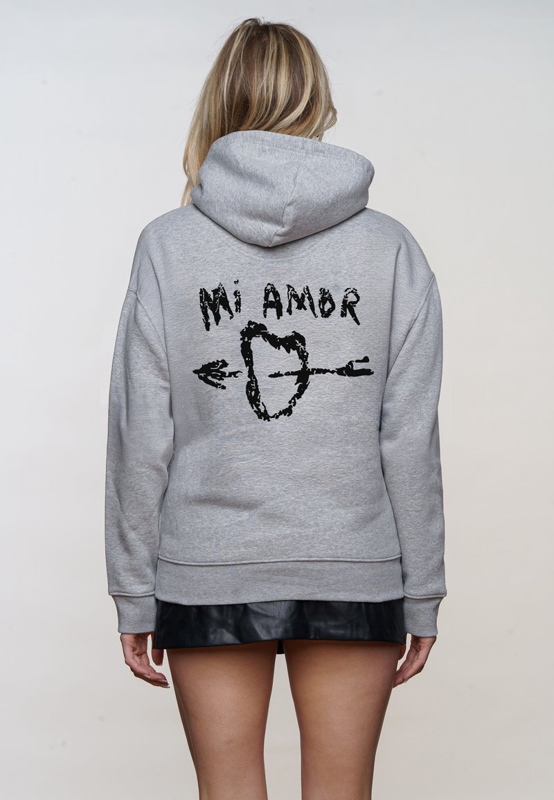 Frau trägt grauen Hoodie mit schwarzem "Mi Amor"-Text und einem Herz, das von einem Pfeil durchbohrt wird, auf der Rückseite, kombiniert mit einem schwarzen Rock.