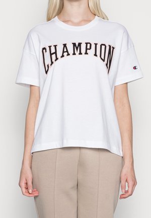 Persona que lleva una camiseta blanca de manga corta con "CHAMPION" en letras grandes negras y rosas en el pecho, y pantalones beige.