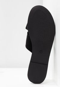 Sandale noire avec une semelle en caoutchouc texturé, un dessus en tissu doux et un design à large bande. Forme plate adaptée à une tenue décontractée.