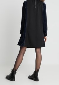 Frau in einem langen schwarzen und marineblauen Samtkleid mit langen Ärmeln und Reißverschluss im Rücken, sheer schwarzen Strumpfhosen und schwarzen geschnürten Stiefeletten steht.