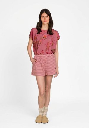 Frau trägt ein rosa geblümtes Kurzarmtop, rosa gestreifte Shorts, beige Socken und flauschige beige Hausschuhe, steht mit einer Hand in der Tasche.