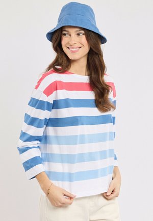 Femme souriante, portant un chapeau seau bleu et une chemise à manches longues avec de larges rayures horizontales rouges, blanches et dans des tons de bleu.
