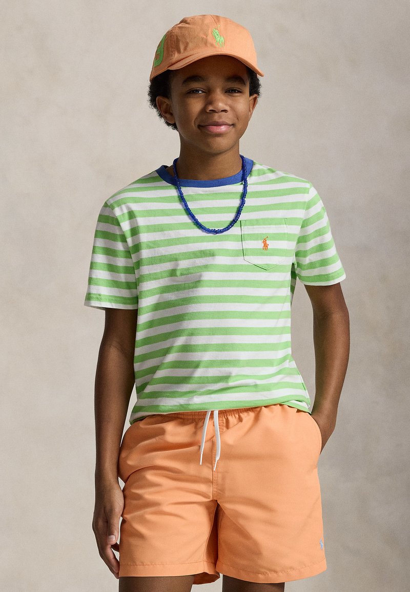 Chlapec s oranžovou čiapkou, zeleno-bielym pruhovaným tričkom, modrým náhrdelníkom z korálok a oranžovými šortkami, ruky vo vreckách.