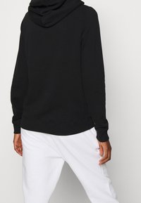 En person i svart hoodie och vita byxor, sedd bakifrån med händerna avslappnat vid sidorna mot en enfärgad bakgrund.