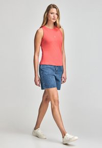 Koronkowy top bez rękawów w kolorze koralowym, niebieskie szorty dżinsowe i białe sneakersy. Stylizacja ma swobodny krój, z prostym logo na topie.