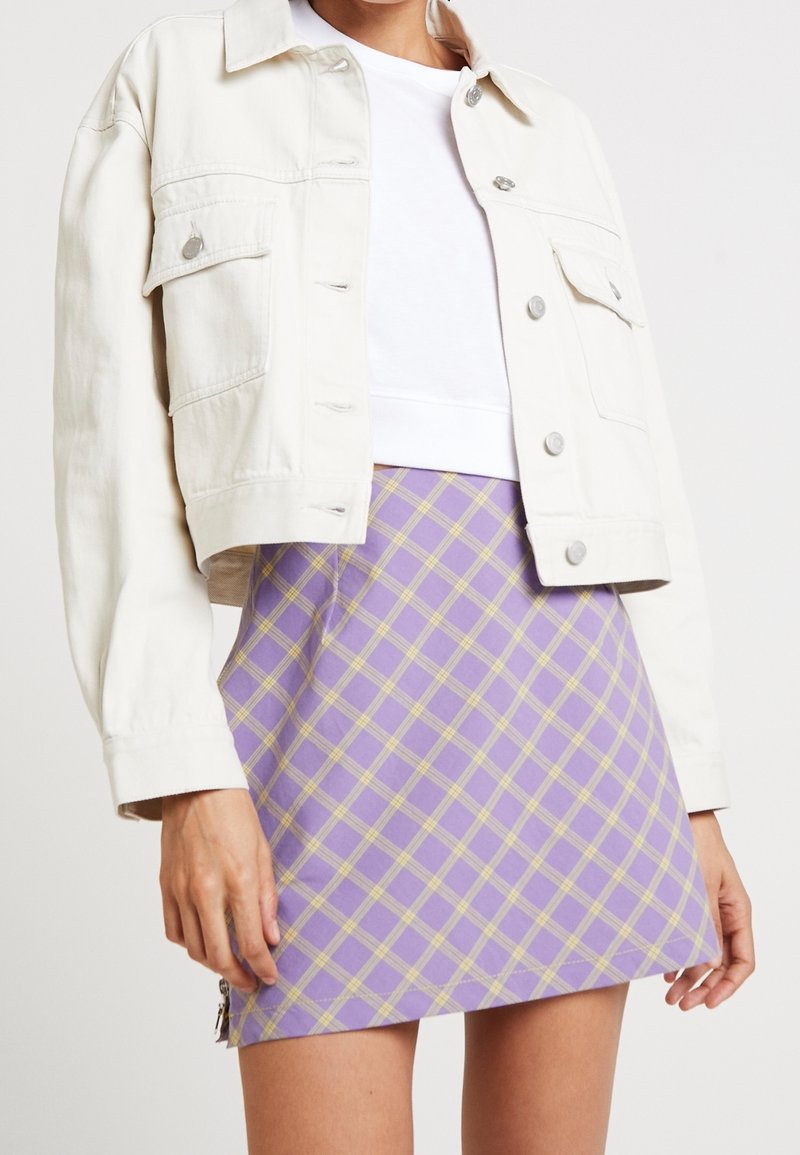 Veste en denim de couleur claire avec fermetures à boutons, portée sur un crop top blanc, associée à une mini jupe à carreaux lilas.
