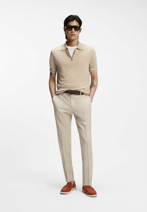 Hombre con camisa beige de manga corta con cremallera, pantalones beige, cinturón marrón, mocasines marrones, gafas de sol negras, de pie con las manos en los bolsillos.